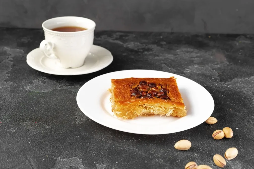 Is baklava healthy? A slice of pistachio baklava on a white plate with a cup of coffee and scattered pistachios on a dark tabletop.